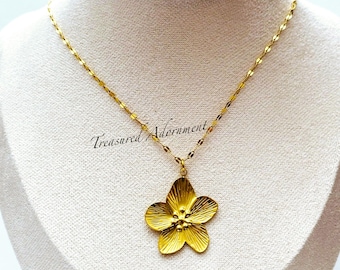 Puede incluir: Un collar dorado con un colgante de flor. El colgante es un diseño detallado de una flor de cinco pétalos. La cadena del collar es delicada y dorada. El texto "Treasured Adornment" es visible.