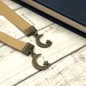 May include: Two beige ribbon bookmarks with bronze crescent moon and star charms. The bookmarks are on a light wood surface. "Treasured Adornment" is written on the surface.