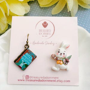 May include: Handmade jewelry set featuring an earring shaped like a book with "Alice's Adventures in Wonderland" and a white rabbit charm holding a pocket watch and a carrot. The jewelry is displayed on a pink card.