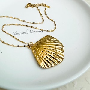 May include: A gold-colored necklace with a large, textured seashell pendant. The pendant has a detailed, ridged surface and is suspended from a delicate chain. The text "Treasured Adornment" is visible on the white surface.