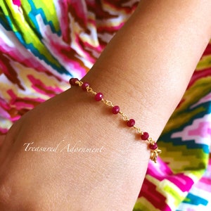 Peut inclure: Un bracelet en chaîne dorée avec de petites perles de pierres précieuses rouge foncé. Le bracelet est au poignet d'une personne.