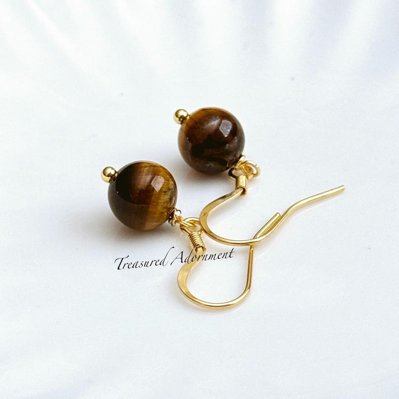 Pu&ograve; includere: Orecchini pendenti color oro con pietre preziose occhio di tigre lucide. Gli orecchini hanno un design a gancio. Il testo "Treasured Adornment" &egrave; visibile. Le pietre misurano circa 1 cm di diametro.