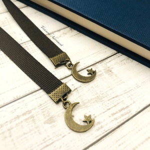 May include: Two brown ribbon bookmarks with gold crescent moon and star charms. The bookmarks are on a white wooden surface.