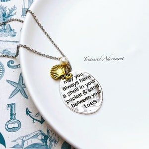 May include: Silver necklace with a gold seashell charm and a silver oval pendant with the text "may you always have a shell in your pocket & sand between your toes".