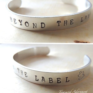Pode incluir: Pulseira de prata com as palavras "Beyond the Label" e uma peça de quebra-cabeça gravada.