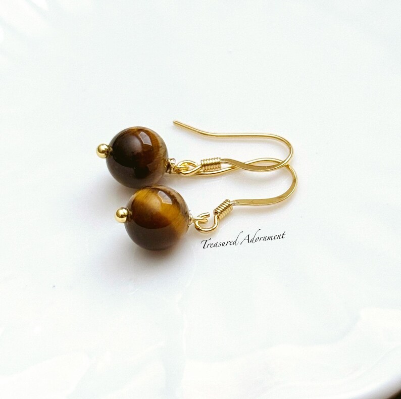 Pu&ograve; includere: Un paio di orecchini color oro con perline rotonde di occhio di tigre marrone. Gli orecchini hanno un semplice design a gancio e il testo "Treasured Adornment" &egrave; visibile.