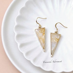 May include: A pair of gold triangle earrings with a white and gold marbled design. The earrings are on a white plate with a scalloped edge.