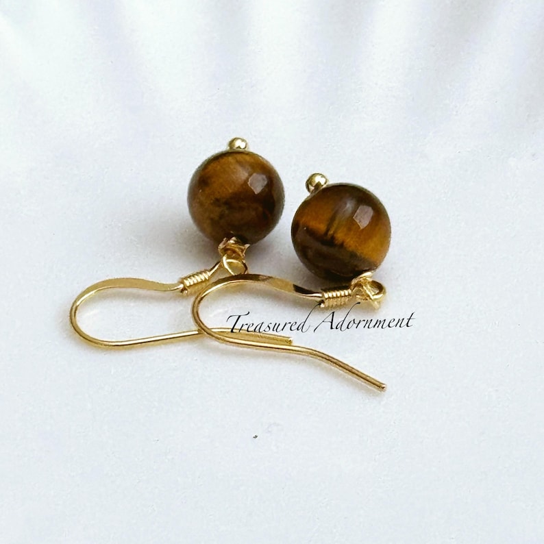 Pu&ograve; includere: Un paio di orecchini color oro con perline rotonde di occhio di tigre marrone. Gli orecchini hanno un design a gancio e il testo "Treasured Adornment" &egrave; visibile. Gli orecchini sono su uno sfondo bianco.