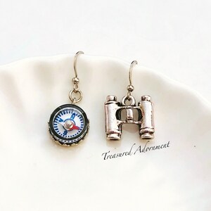 Puede incluir: Un par de pendientes de plata con un dije de brújula y binoculares. La brújula tiene una cara azul y blanca con una aguja roja. Los binoculares son de plata y tienen un diseño detallado. Los pendientes cuelgan de ganchos de plata.