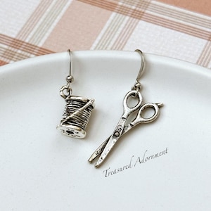 May include: A pair of silver earrings with a spool of thread and a pair of scissors charms. The earrings are on a white background. "Treasures Adornment" is written in the bottom right corner of the image.