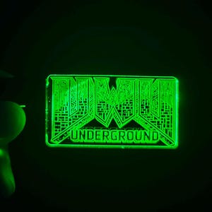 以下が含まれることがあります： 幾何学模様と「UNDERGROUND」の文字が入った、緑色に光る長方形のピン。暗い背景の中で、明るい緑色の光を放っています。