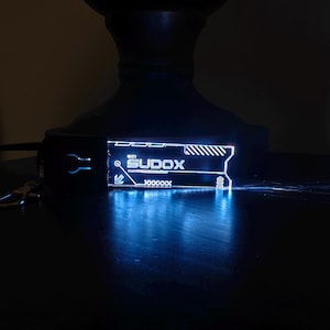 May include: A glowing blue keychain with the word "SUDOX" in white, along with other white graphic elements. The keychain is attached to a black strap with a silver clasp. The design is illuminated, creating a futuristic aesthetic.