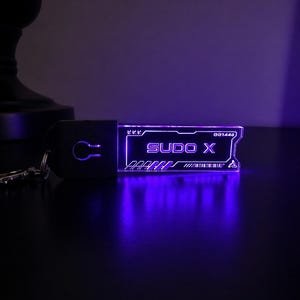 May include: A glowing purple keychain with the text "SUDO X" and a key symbol. The keychain has a rectangular design with a futuristic aesthetic. The background is dark, highlighting the illuminated details.