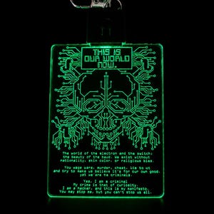 Op de afbeelding: Een rechthoekige acryl sleutelhanger die felgroen oplicht, met een print van een schedel in printplaat-stijl en de tekst: THIS IS OUR WORLD NOW. De tekst van het Hacker Manifesto staat onder de schedel, inclusief een zilveren metalen karabijnhaak.