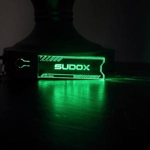May include: A glowing green keychain with the word "SUDOX" in white, featuring a futuristic design. The keychain is attached to a black holder and a silver clasp. The image is dark, with the green light illuminating the keychain.