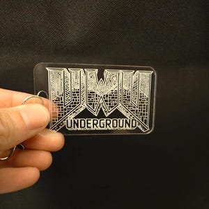 以下が含まれることがあります： 「UNDERGROUND」の文字と白いスタイリッシュなロゴが刻まれた、透明な長方形のキーホルダー。キーホルダーには、鍵を取り付けるためのシルバー色のリングが付いています。
