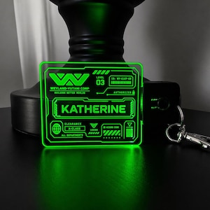 Könnte beinhalten: Ein leuchtend grünes Acryl-ID-Abzeichen mit dem Text "WEYLAND-YUTANI CORP" und dem Namen "KATHERINE". Das Abzeichen hat ein futuristisches Design mit verschiedenen Symbolen und einem Schlüsselanhänger. Das Abzeichen ist beleuchtet und erzeugt ein helles grünes Leuchten.