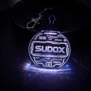 May include: A glowing, circular keyring featuring the word "SUDOX" in a futuristic font. The design incorporates circuit board patterns and a keyhole symbol. The keyring is attached to a black strap and a silver clasp.