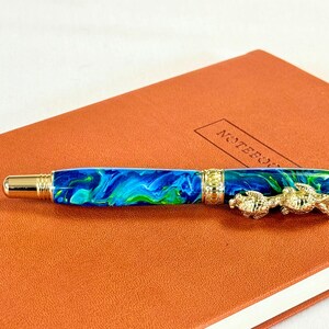 The Sea Turtle Roller Ball pen features a stunning blue-ocean synthetic Fire Opal, turtle shell texture, and sea turtle clip. Truly a turtle lover gift. It comes either as a Rollerball or Fountain Pen. The cap posts securely with wide threads that won't cross. Hawaiian Honu Pen that has beautiful ocean themed features. One of my favorite Sea Creatures is the Sea Turtle. This Pen has it all. The pen comes with a windowed pen box making this item gift-ready.