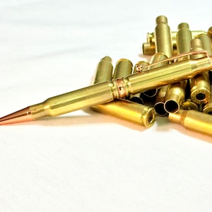 Op de afbeelding: Een balpen gemaakt van een koperen geweerpatroonhuls met een koperkleurige punt en een metalen zakclip, geplaatst voor een kleine stapel lege koperen kogelhulzen op een effen witte ondergrond.