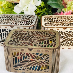 Peut inclure: Trois boîtes en bois avec des motifs de feuilles complexes. Les boîtes sont en bois clair et ont une finition naturelle. La boîte du milieu est ouverte et remplie de bonbons colorés.