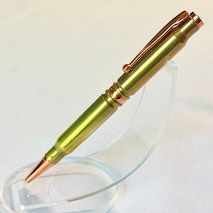 Op de afbeelding: Een balpen ontworpen als een geweerpatroon, met een goudkleurige metalen behuizing, een koperkleurige punt en een bijpassende koperkleurige pocketclip. De pen wordt gepresenteerd op een gebogen standaard van transparant acryl.