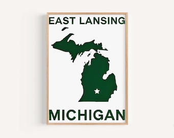 Könnte beinhalten: Ein weißer gerahmter Druck mit dem Text "EAST LANSING MICHIGAN" in grünen Buchstaben. Der Druck zeigt eine grüne Silhouette des Bundesstaates Michigan mit einem weißen Stern in der Mitte.