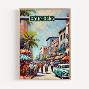 Op de afbeelding: Een ingelijste kunstdruk van Calle Ocho, een levendige straatscène met kleurrijke gebouwen, palmbomen en mensen. Een groen straatnaambord leest "Calle Ocho". Een vintage turquoise auto staat rechts.