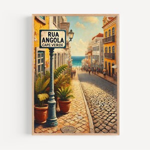 Puede incluir: Una impresión enmarcada de una escena callejera en Cabo Verde, con una calle adoquinada que conduce al océano. Un letrero de la calle dice "Rua Angola Cape Verde". Los edificios con fachadas amarillas y blancas bordean la calle, y las plantas en macetas están en la base de una farola.