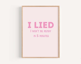 May include: A framed pink art print with the words "I LIED" in large, bubble-style letters, followed by "I WON'T BE READY IN 5 MINUTES" in a smaller font. The frame is a light wood color.