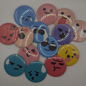 Puede incluir: Colección de botones redondos con emojis coloridos. Los botones son azules, rosas, rojos, amarillos y morados. Cada botón muestra una expresión facial diferente, como enfado, tristeza o alegría. Un accesorio divertido y expresivo.