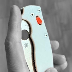 Peut inclure: Un couteau de poche blanc avec un logo d'araignée noir et un bouton rouge sur le manche.