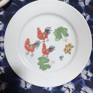 May include: A white ceramic plate with a red, black, and green rooster design. The plate features three roosters, green leaves, and yellow flowers.
