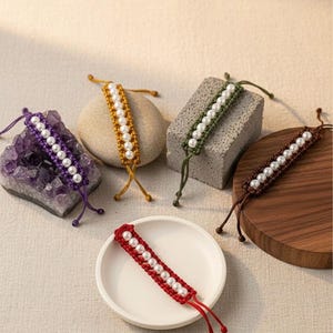 May include: A collection of handmade macrame bracelets in various colors, including purple, gold, green, brown, and red. Each bracelet features a row of white pearls. The bracelets are displayed on a variety of surfaces, including a stone, a wooden disc, and a white ceramic dish.