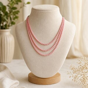 May include: A three-strand pink pearl necklace displayed on a beige mannequin bust. The necklace is composed of small, round pearls. The bust is mounted on a wooden base, and the background includes a vase with greenery and white flowers.