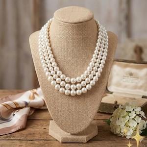May include: A three-strand pearl necklace displayed on a tan jewelry bust. The pearls are white and evenly sized. The bust is on a wooden surface, with a vintage jewelry box and a floral arrangement in the background.