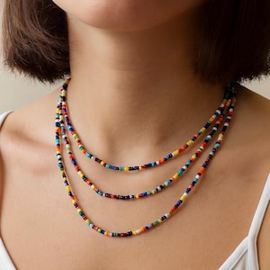 May include: A multi-strand necklace featuring small, colorful beads in a variety of colors including red, orange, yellow, green, blue, and white. The necklace is layered and worn around the neck, creating a vibrant and eye-catching accessory.