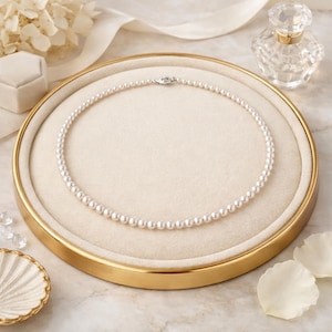 May include: A delicate pearl necklace with a silver clasp rests on a gold-rimmed tray with a cream-colored interior. The necklace is composed of numerous small, round, white pearls. The tray is surrounded by decorative elements, including a small perfume bottle and flower petals.