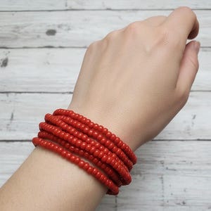 May include: A stack of six red beaded bracelets encircles a wrist. The bracelets are made of small, round beads, and the color is a vibrant, solid red. The background is a light-colored, wooden surface.