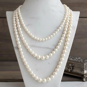 May include: Three strands of white pearl necklaces displayed on a white marble jewelry bust. The pearls vary in size, with the largest at the bottom. A small, ornate silver box is visible in the background.