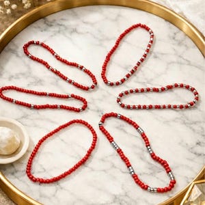 May include: Six red beaded necklaces with silver accents are arranged on a marble-patterned surface within a gold-rimmed tray. The necklaces are composed of small, round red beads interspersed with sections of silver beads. The tray is surrounded by decorative elements.