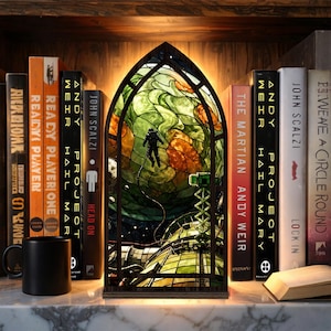 May include: A stained glass lamp with a space-themed design. The lamp features an astronaut floating in space, with green, orange, and blue colors. The lamp is set on a shelf with books and a black mug.