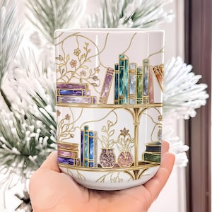 Pode incluir: Um copo cil&iacute;ndrico branco com uma ilustra&ccedil;&atilde;o envolvente de duas prateleiras cheias de livros coloridos e plantas em vasos. O design utiliza linhas douradas para criar um efeito de vitral, exibindo lombadas de livros em tons roxos, azuis e verdes.