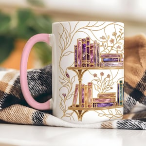 Pode incluir: Uma caneca de cer&acirc;mica branca com uma asa rosa pastel, apresentando uma ilustra&ccedil;&atilde;o de linhas douradas de duas prateleiras cheias de livros com aspeto de cristal em tons de roxo, rosa e azul, com delicadas vinhas douradas.