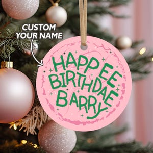May include: A round, pink Christmas ornament with the text "HAPPY BIRTHDAE BARRY" in green. The ornament has a light pink background with a decorative border and a tan ribbon for hanging. The ornament is hanging on a Christmas tree.