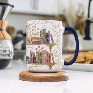 Pode incluir: Uma caneca de cer&acirc;mica branca com uma asa azul-escura, apresentando uma ilustra&ccedil;&atilde;o com linhas douradas de duas prateleiras cheias de livros coloridos e detalhes florais. O design inclui v&aacute;rias lombadas de livros em tons de roxo, verde e castanho.