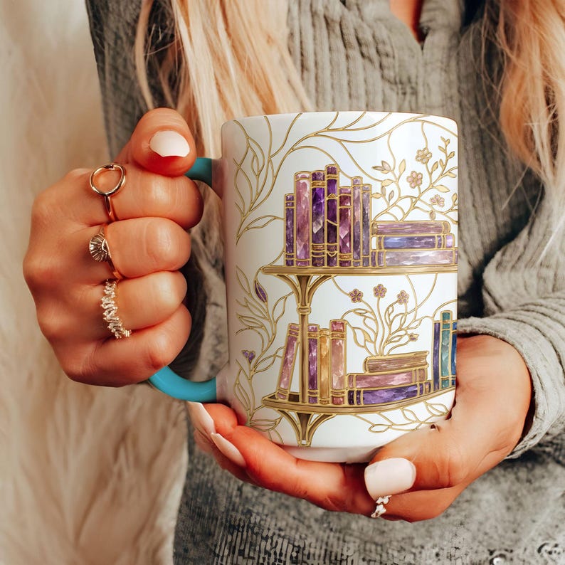 Pode incluir: Uma caneca de cer&acirc;mica branca com uma asa azul-petr&oacute;leo, apresentando uma ilustra&ccedil;&atilde;o com contornos dourados de duas prateleiras cheias de livros roxos e cor-de-rosa, acentuada por delicadas vinhas florais douradas na superf&iacute;cie exterior.