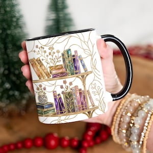 Pode incluir: Uma caneca de cer&acirc;mica branca com asa e rebordo pretos, apresentando uma ilustra&ccedil;&atilde;o detalhada de duas prateleiras cheias de livros coloridos, pequenas plantas em vasos e delicadas linhas douradas de videiras em toda a superf&iacute;cie.