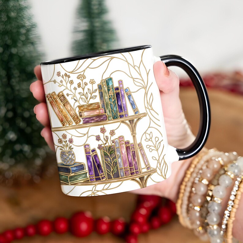 Pode incluir: Uma caneca de cer&acirc;mica branca com asa e rebordo pretos, apresentando uma ilustra&ccedil;&atilde;o detalhada de duas prateleiras cheias de livros coloridos, pequenas plantas em vasos e delicadas linhas douradas de videiras em toda a superf&iacute;cie.