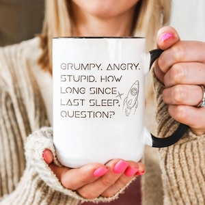 May include: A white ceramic mug with a black handle and interior. The front features black text that reads: GRUMPY. ANGRY. STUPID. HOW LONG SINCE LAST SLEEP, QUESTION? Next to the text is a small black line drawing of a rocket ship with a star.